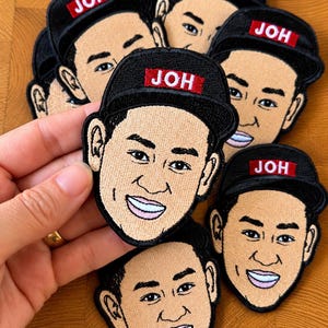May include: A collection of embroidered patches featuring a cartoon face wearing a black baseball cap with a red rectangular label that has the white text JOH printed on it. The face has tan skin, dark hair, and a wide smile with visible teeth.