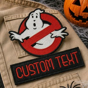 Può includere: Una toppa ricamata circolare raffigurante un personaggio fantasma bianco all'interno di un cerchio rosso con una barra diagonale, posizionata sopra una toppa rettangolare nera con il testo ricamato in rosso CUSTOM TEXT su tessuto beige.
