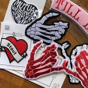 May include: A collection of embroidered patches with various designs. One patch is heart-shaped with the words "MUTUAL WEIRDNESS FOREVER". Another features a red heart with a banner that reads "Stout". Other patches depict skeleton hands.