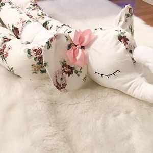 Puede incluir: Una almohada blanca con forma de elefante con tela de estampado floral y un lazo rosa. La almohada está sobre una superficie blanca y esponjosa.