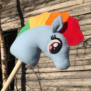 Pode incluir: Uma cabeça de cavalo de pelúcia azul e arco-íris em um bastão de madeira. O cavalo tem um olho preto com uma pupila rosa e branca e um cílio preto.