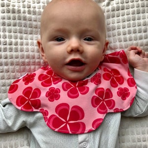 Puede incluir: Un bebé con un babero rosa y rojo con un patrón floral. El babero tiene un borde festoneado.