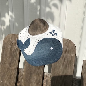 Puede incluir: Un babero blanco con un diseño de ballena azul. La ballena está hecha de mezclilla y tiene un botón blanco en su vientre. El babero tiene lunares blancos sobre un fondo blanco.