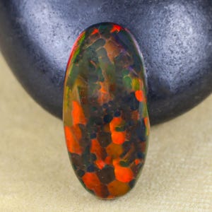 May include: An oval-shaped gemstone with a vibrant display of colors. The stone features a mix of orange, green, and black hues, creating a unique pattern. The gemstone is set against a neutral background and a dark, smooth stone.