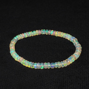 May include: A delicate bracelet composed of small, iridescent opal beads. The beads display a range of colors, including green, blue, and yellow, and are strung together to form a circular shape. The bracelet is set against a dark background.