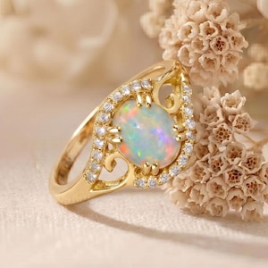 May include: A gold ring featuring an oval opal gemstone with iridescent colors, surrounded by small, clear gemstones. The ring has a decorative, openwork design on the sides.