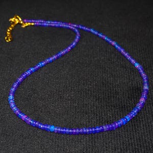 May include: A delicate necklace composed of small, round, iridescent beads in shades of blue, purple, and pink. The necklace has a gold-colored clasp and chain. The beads are strung together to form a choker-style necklace.