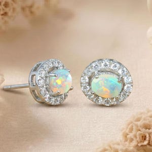 May include: A pair of stud earrings featuring oval-shaped opals surrounded by a halo of small, clear gemstones. The opals display a play of color, with flashes of blue, green, and yellow. The earrings are set in a silver-colored metal.