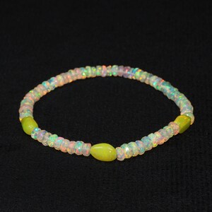 May include: A delicate bracelet featuring iridescent opal beads interspersed with three oval-shaped, light green beads. The bracelet is strung on a flexible band and is displayed against a dark background.