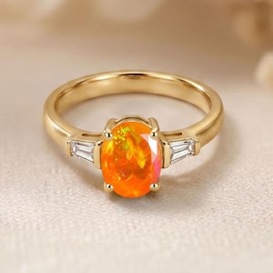 May include: A gold ring featuring an oval-cut, fiery orange opal gemstone as the centerpiece. Two baguette-cut white gemstones flank the opal. The ring has a polished gold band and is set against a neutral background.