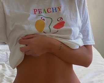 Op de afbeelding: Wit T-shirt met het woord "PEACHY" bedrukt in regenboogkleuren. Het shirt heeft een kleurrijk abstract gezichtontwerp. Het woord "EMIC" is getatoeëerd op de pols van de persoon.