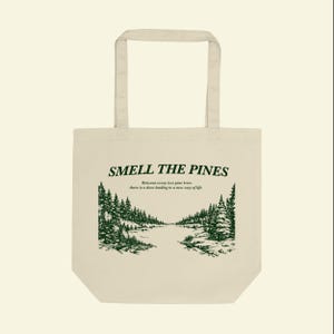 May include: Cream-colored tote bag with the text "SMELL THE PINES" in dark green. Below the text is a green illustration of a forest scene. The bag has two handles.