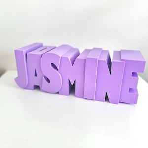 May include: A lavender-colored, three-dimensional word "JASMINE" is displayed on a white surface. The letters are blocky and stand upright, creating a visually striking name display. The background is a plain white wall.