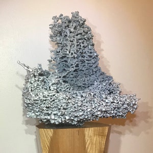 May include: A silver aluminum sculpture resembling a honeycomb structure, displayed on a wooden pedestal with a natural wood grain.