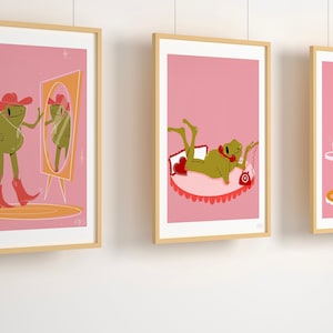 set of 3 pink frog art prints in wooden frames. Frog wears cowboy boots and cowgirl hat in pink