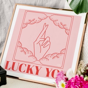 square gold frame rests against a white sofa surrounded by house plants. Square art is pink and red and inspired by retro tarot art. At the bottom of the art, retro red typography reads LUCKY YOU