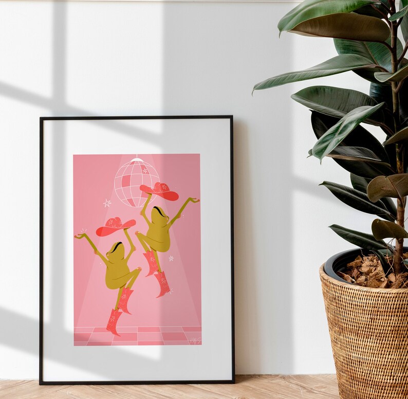 pink dancing frog art print rests against a white wall bathed in sunlight next to a tall house plant. Frogs wear cowboy hat and boots in a sparkling hot pink colour