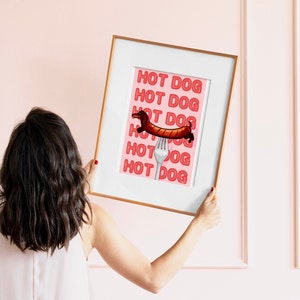 Printable pink & red dog art print. Soft pink background with red text in the style of neon lights reading HOT DOG. in the centre of the art print is a dachshund in the shape of a sausage on the end of a silver fork in a playful style in wood frame