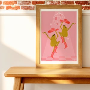 pink dancing frog art print rests against a wooden side board in front of a brick wall
