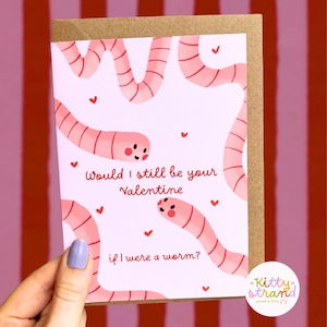 worm Valentine's Day card. pink Valentine's day card with an illustration of two worms surrounded by love hearts and text reading Would I still be your valentine if I were a worm? in red typography. Valentine's Day card for boyfriend, husband or partner held by a woman in front of a cosy background