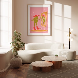 pink and green cowboy frog art print of a cartoon style frog wearing a pink cowgirl hat and pink cowboy boots looking into a vintage yellow mirror. Print features a soft pink background and sits in a gold frame hanging above a white sofa