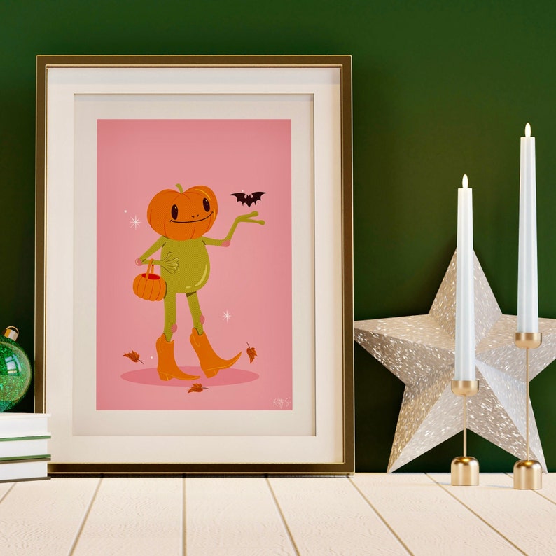 howdy halloween frog art print in pink, green and orange next to candles. UPS style cartoon illustration of a frog with a pumpkin head wearing orange cowboy boots and carrying a pumpkin bag. next to him flies a little bat, leaves surround his feet