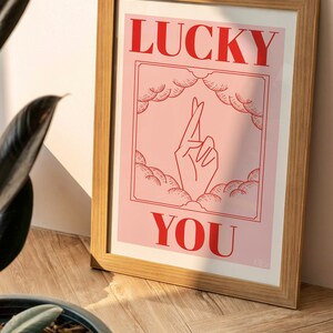 wooden frame with pink and red Lucky You art print resting against a living room wall bathed in sunlight and shadows