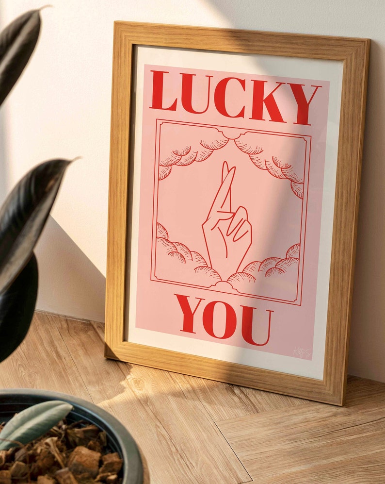 wooden frame with pink and red Lucky You art print resting against a living room wall bathed in sunlight and shadows
