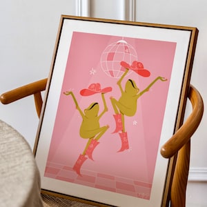 wooden mid-century frame sits in a wooden dining room chair. Art in frame is pink and features two dancing frogs in pink sparkly cow girl boots and hats beneath a disco ball on a pink dance floor