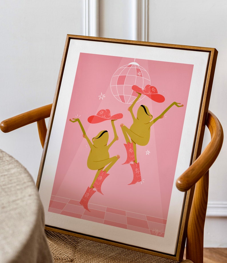 wooden mid-century frame sits in a wooden dining room chair. Art in frame is pink and features two dancing frogs in pink sparkly cow girl boots and hats beneath a disco ball on a pink dance floor