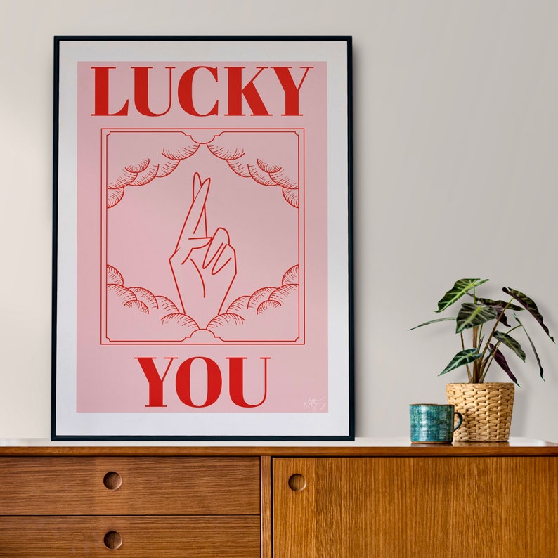 A3 pink and red Lucky art print sits on a mid-century side board next to a potted plant and mug of coffee