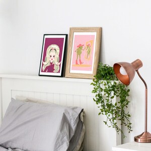 Two frames rest against a headboard next to a green plant and an iron lamp. First frame has a vintage style art print of a woman with green 60s style hair. The second frame features a retro style frog wearing pink cowgirl boots & a pink cowgirl hat