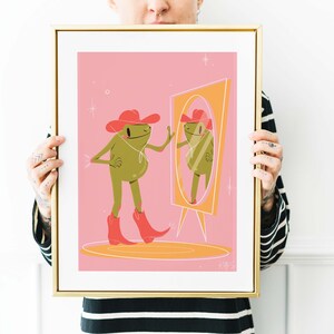 Women with tattoos holding up a golden frame. Art in frame features a UPA style frog illustration. A Green smiling frog looks into a tall yellow mirror wearing pink cowgirl boots and a pink cowgirl hat