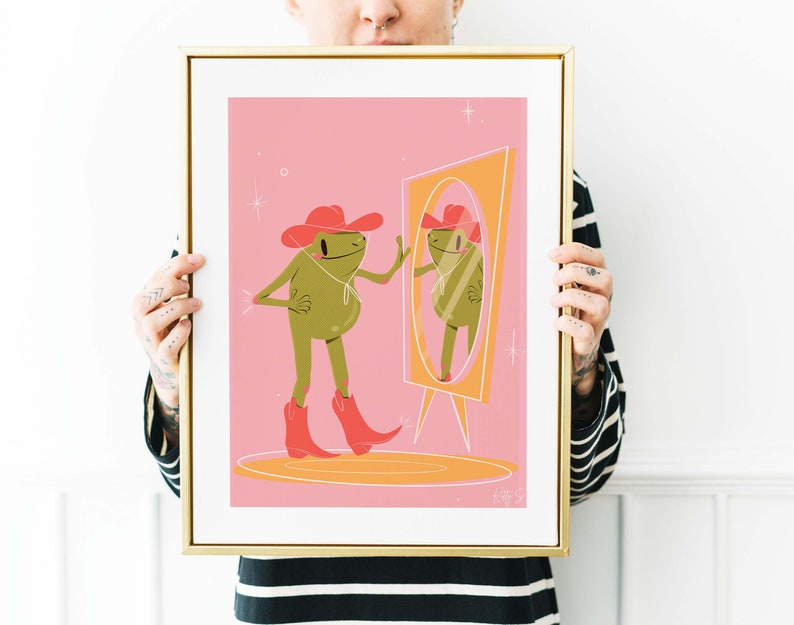 Women with tattoos holding up a golden frame. Art in frame features a UPA style frog illustration. A Green smiling frog looks into a tall yellow mirror wearing pink cowgirl boots and a pink cowgirl hat
