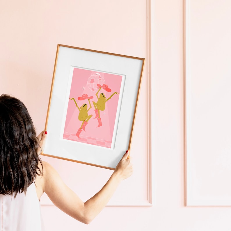 women holds a pink frog art print in a wooden frame as she hangs it on a pink bedroom wall. Print features two frogs in cow girls hats and boots dancing beneath a pink disco ball