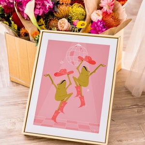 Gold frame leans against a box of flowers. In the frame is a pink art print of 2 green frogs wearing cow girl hats and boots dancing beneath a sparkling disco ball on a pink dance floor surrounded by hand drawn sparkles