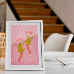 pink frog art print of two dancing frogs in cow girl hats and boots dancing beneath a disco ball. Print sits in a white frame on a dining room table