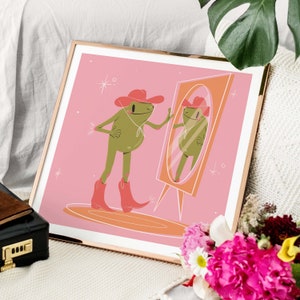 Gold frame leans against white sofa surrounded by flowers. Art in frame features a UPA style frog illustration. A Green smiling frog looks into a tall yellow mirror wearing pink cowgirl boots and a pink cowgirl hat