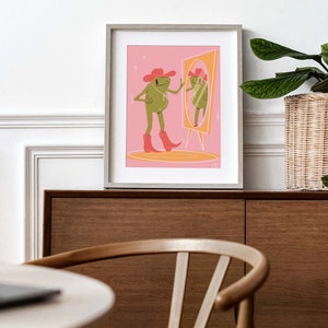 On a retro sideboard a wooden frame and green house plant sit. Art print in the frame features a UPA style frog illustration. A Green smiling frog looks into a tall yellow mirror wearing pink cowgirl boots and a pink cowgirl hat