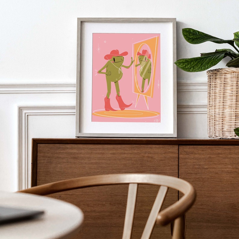 On a retro sideboard a wooden frame and green house plant sit. Art print in the frame features a UPA style frog illustration. A Green smiling frog looks into a tall yellow mirror wearing pink cowgirl boots and a pink cowgirl hat