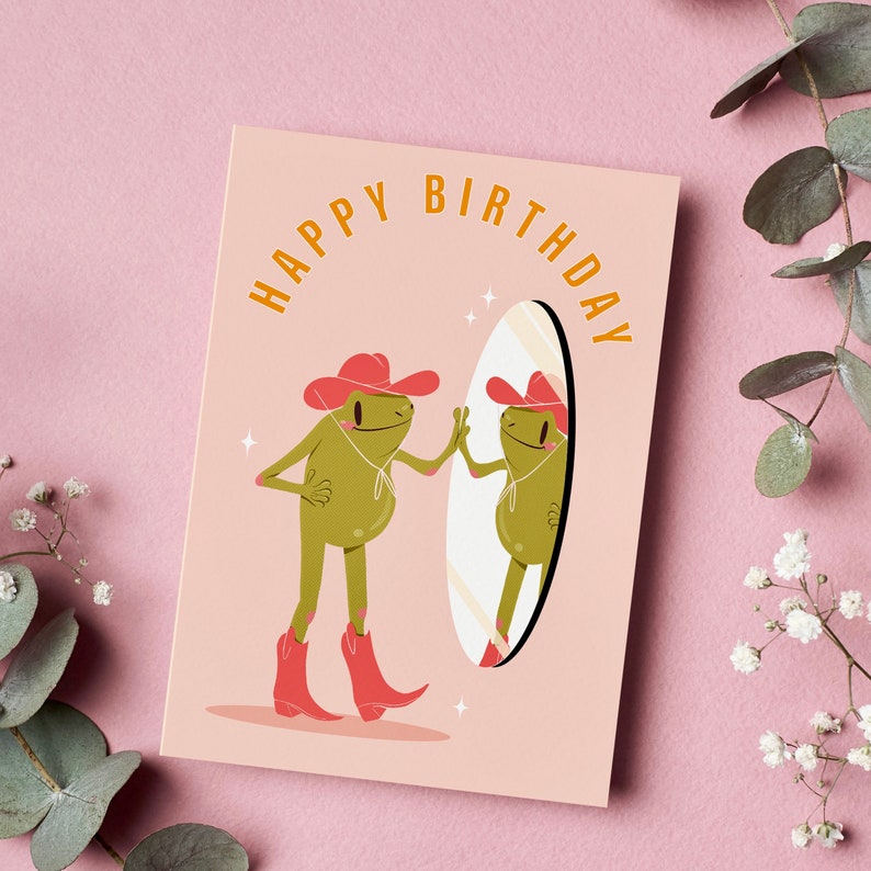 Pink and green cowboy frog happy birthday card. pink greetings card with a green frog in a cowboy hat and cowboy boots looking I the mirror and smiling. text on the card reads happy birthday in yellow lettering. Card rests against a pink background