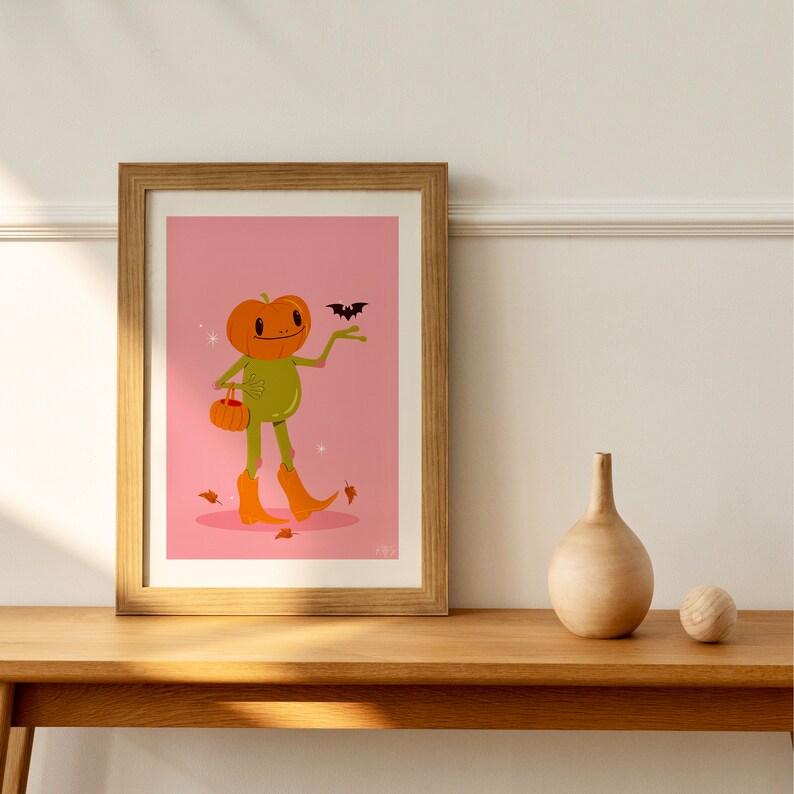 howdy halloween frog art print in pink, green and orange on wooden side. UPS style cartoon illustration of a frog with a pumpkin head wearing orange cowboy boots and carrying a pumpkin bag. next to him flies a little bat, leaves surround his feet