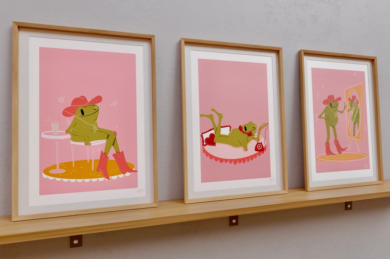 set of 3 pink frog art prints in wooden frames. Frog wears cowboy boots and cowgirl hat in pink