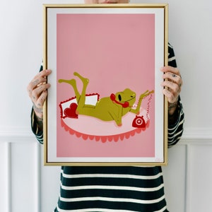 Women with tattoos holding up a golden frame. Art in frame features a UPA style frog illustration. A Green smiling whilst laid on a pink and red valentines aesthetic bed talking on a retro phone