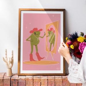 Wooden frame sits next to a bunch of flowers on top of a wall. A women stands to the right. Frame features a UPA style frog illustration. A Green smiling frog looks into a tall yellow mirror wearing pink cowgirl boots and a pink cowgirl hat