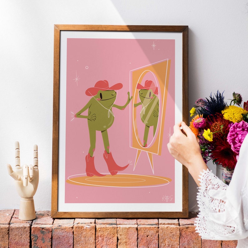 Wooden frame sits next to a bunch of flowers on top of a wall. A women stands to the right. Frame features a UPA style frog illustration. A Green smiling frog looks into a tall yellow mirror wearing pink cowgirl boots and a pink cowgirl hat