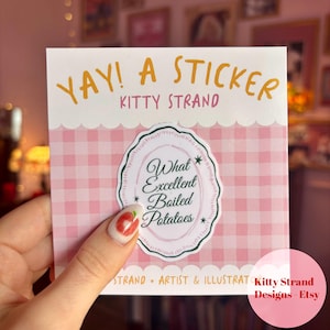 Op de afbeelding: Een vierkante sticker met een roze ruitjespatroon. De sticker heeft de tekst "YAY! A STICKER" in geel en "KITTY STRAND" in een kleiner lettertype. Een centraal ovaal ontwerp luidt "What Excellent Boiled Potatoes."