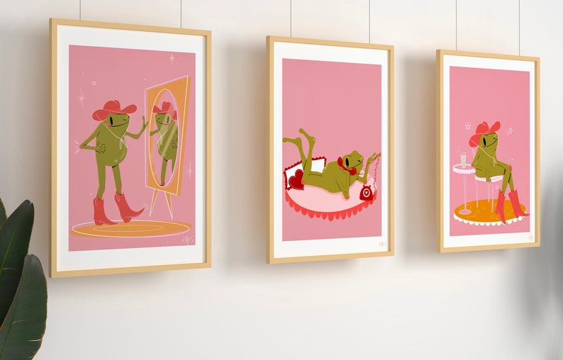 set of 3 pink frog art prints in wooden frames. Frog wears cowboy boots and cowgirl hat in pink