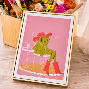 gold frame with pink and green cow boy frog art print. Poster shows a green frog wearing pink cow boy boots and pink hat sitting at a table drinking a little iced coffee. Frame rests against box of fresh flowers