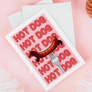 Printable pink & red dog art print. Soft pink background with red text in the style of neon lights reading HOT DOG. in the centre of the art print is a dachshund in the shape of a sausage on the end of a silver fork in a playful style on pink table